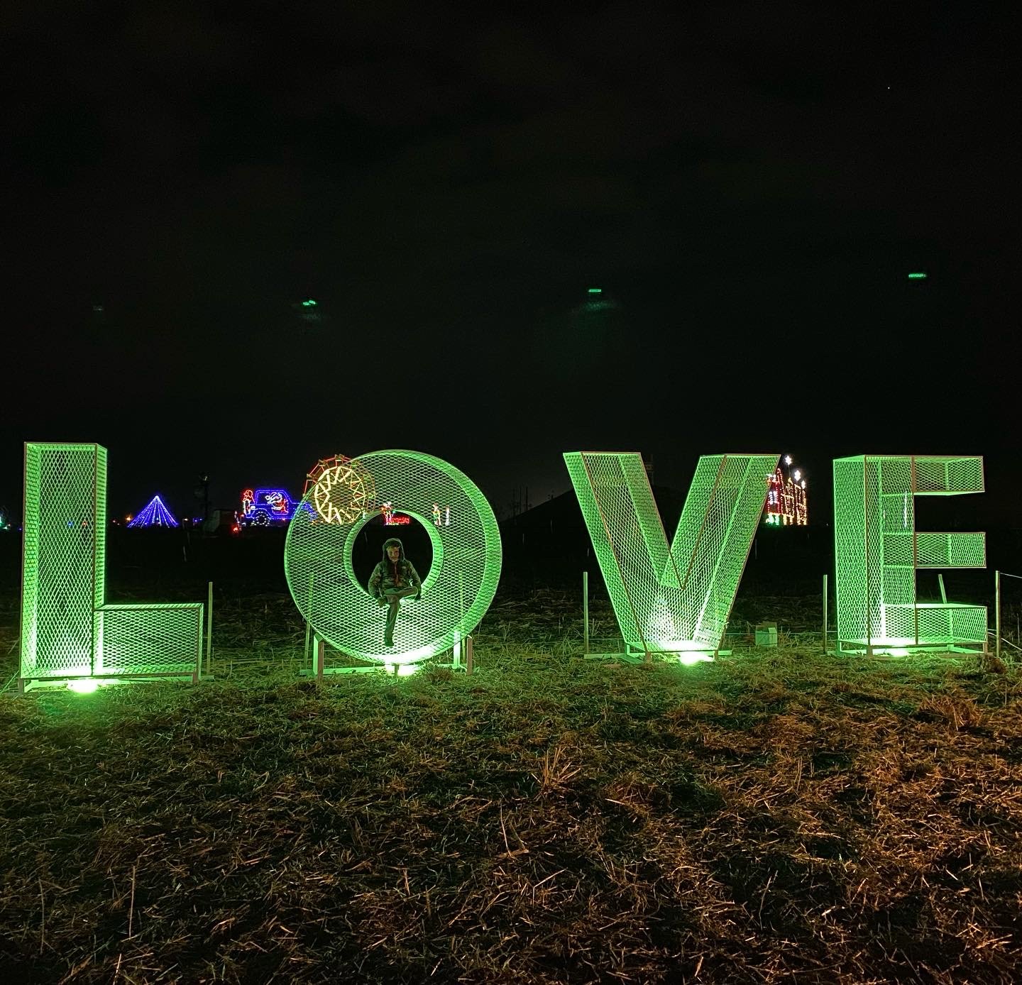 44F520C0-45AB-4372-BE40-A327102DD73D Large illuminated green "LOVE" sign at night, with a person sitting inside the "O," surrounded by festive lights and decorations in the background.