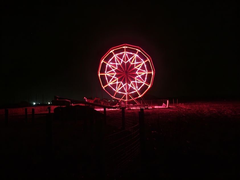 6CA4A5DD-7D01-4878-A63B-D67D675DC4FF Illuminated red ferris wheel against a dark night sky, showcasing vibrant neon lights in a rural setting.