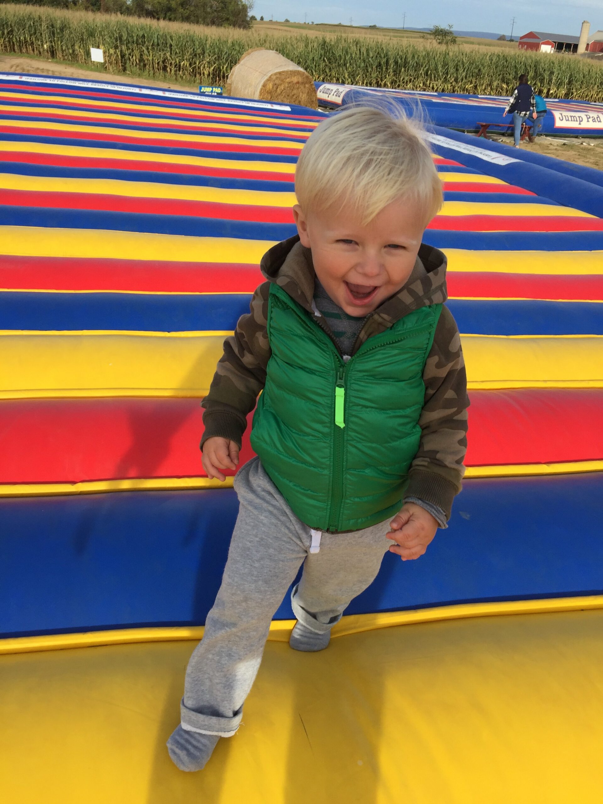 Country+creek+produce+farm++Pa+-+94DABC79-4394-4570-99DE-0214369CB2E0 Smiling toddler wearing a green vest and gray pants playing on a colorful inflatable jump pad at a farm, surrounded by cornfields and hay bales.