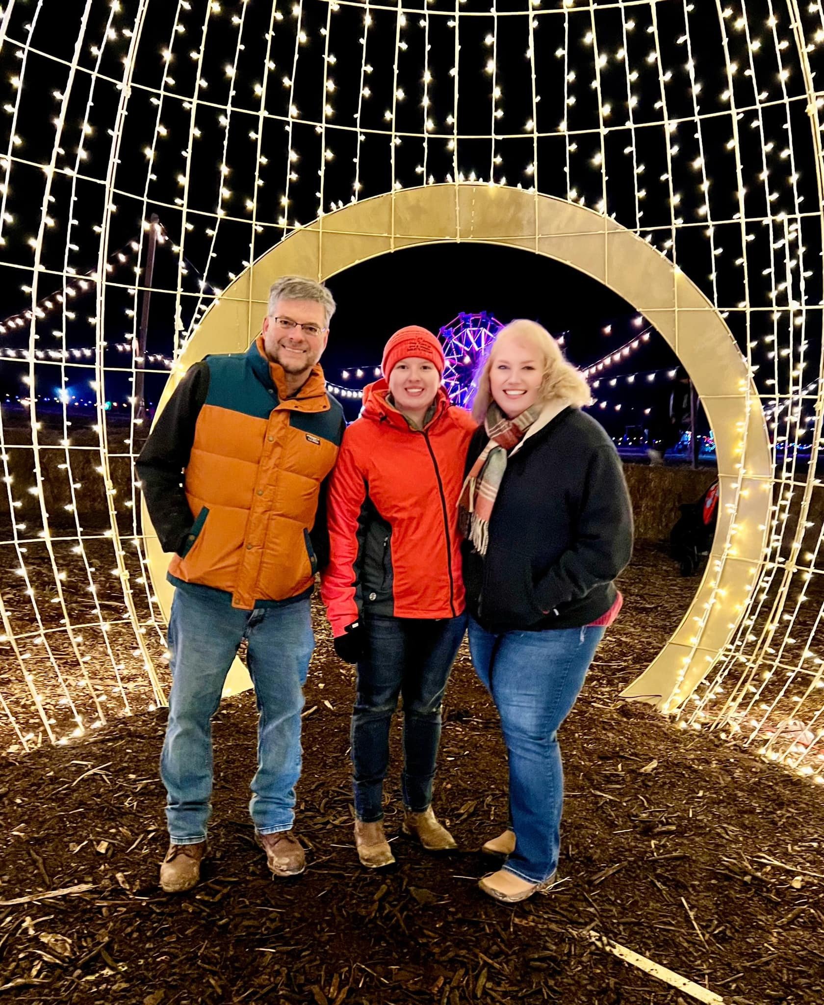 IMG_8615 Three friends smiling together in front of a festive light display at night, surrounded by decorative lights and a golden arch.