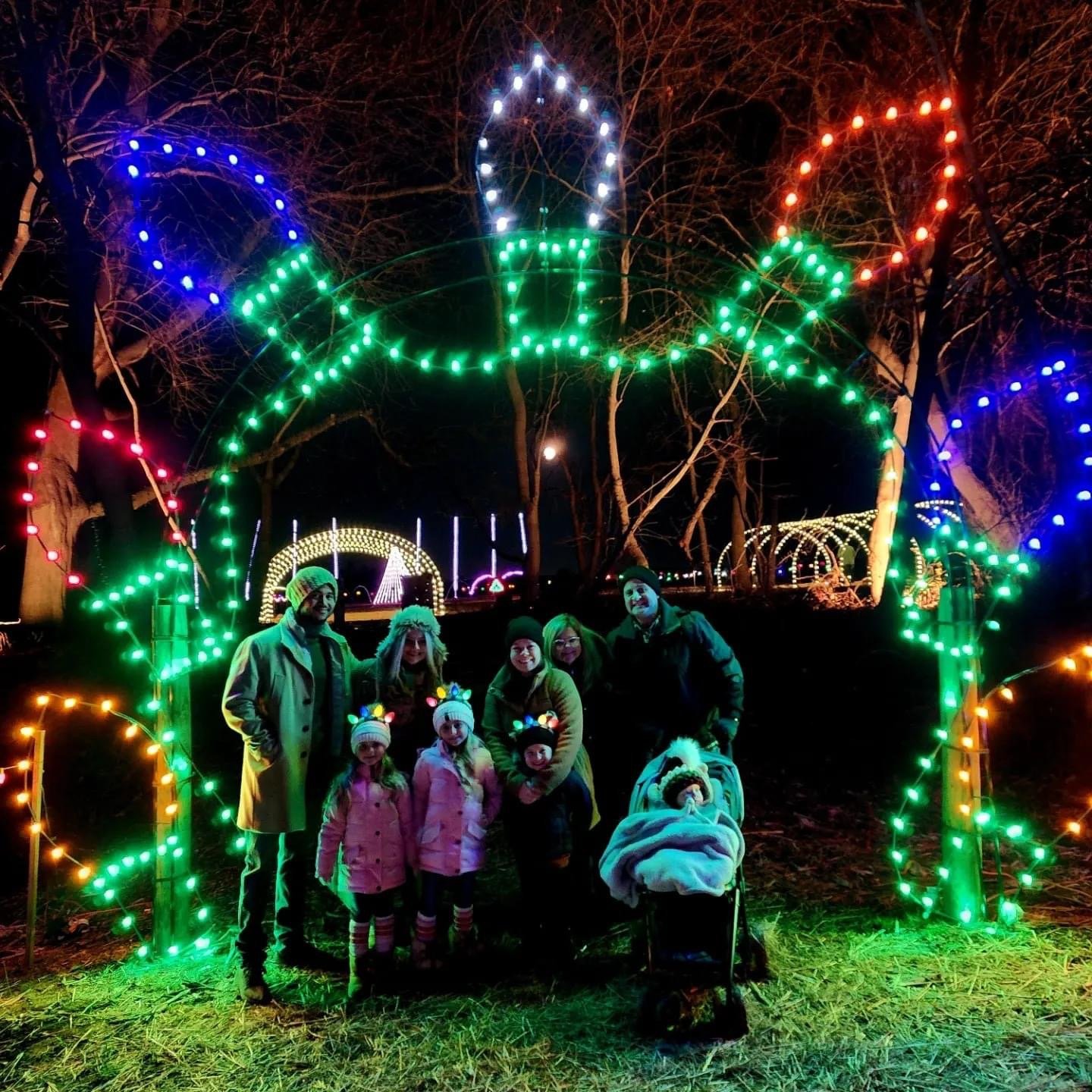 IMG_8616 Family posing in front of colorful holiday lights shaped like oversized Christmas bulbs at a festive outdoor display during the evening, surrounded by illuminated decorations and trees.