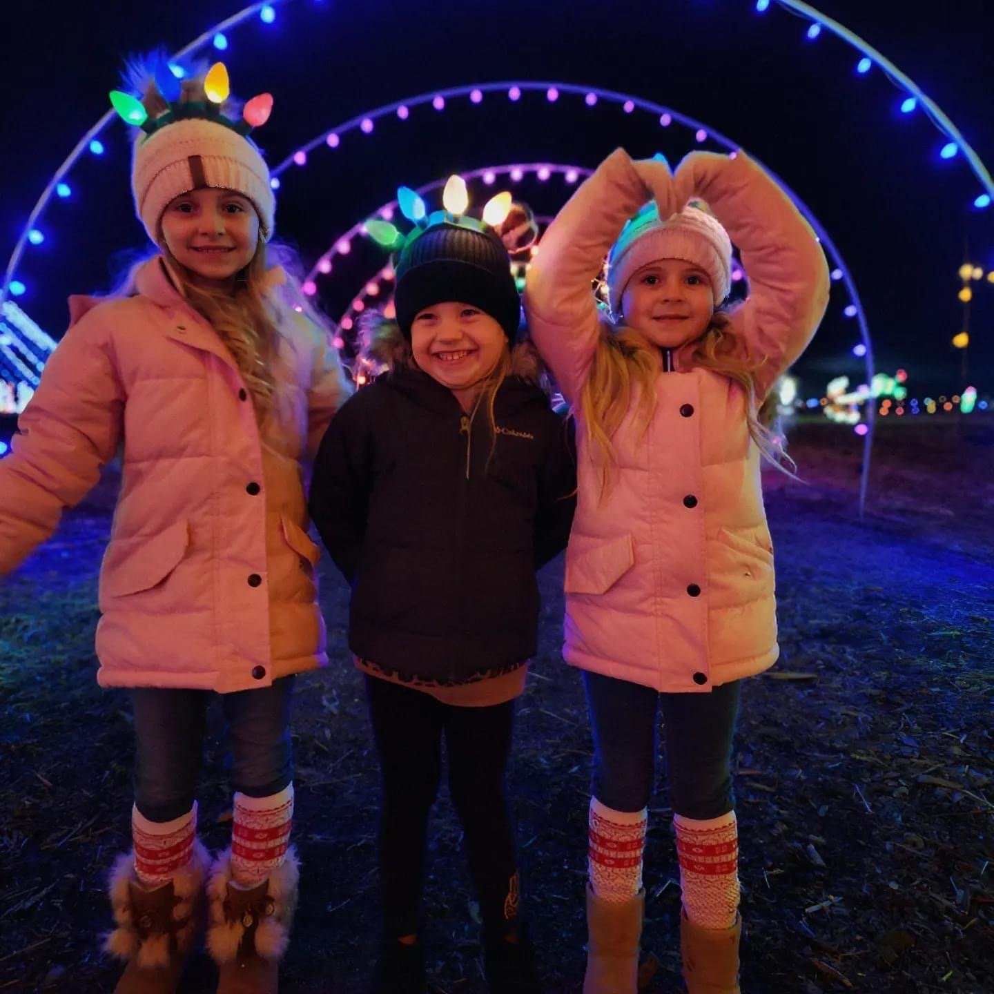 IMG_8617 Three children smiling and posing in winter clothing at a colorful holiday light display, with vibrant lights illuminating the background. The scene captures a festive atmosphere, perfect for family holiday activities.