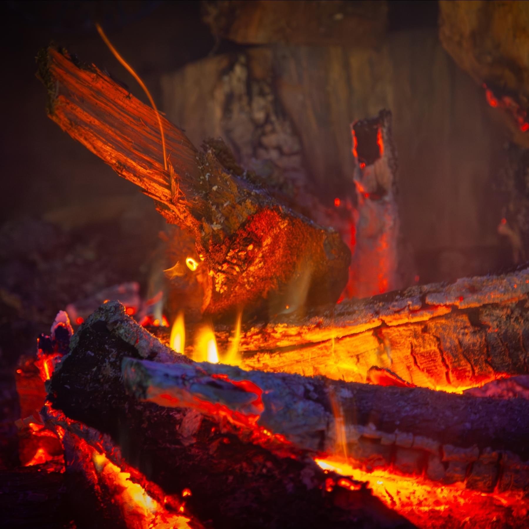 IMG_9747 Close-up of glowing embers and burning logs in a fireplace, showcasing vibrant orange and red flames against dark, charred wood. Ideal for illustrating cozy atmospheres or fire safety topics.