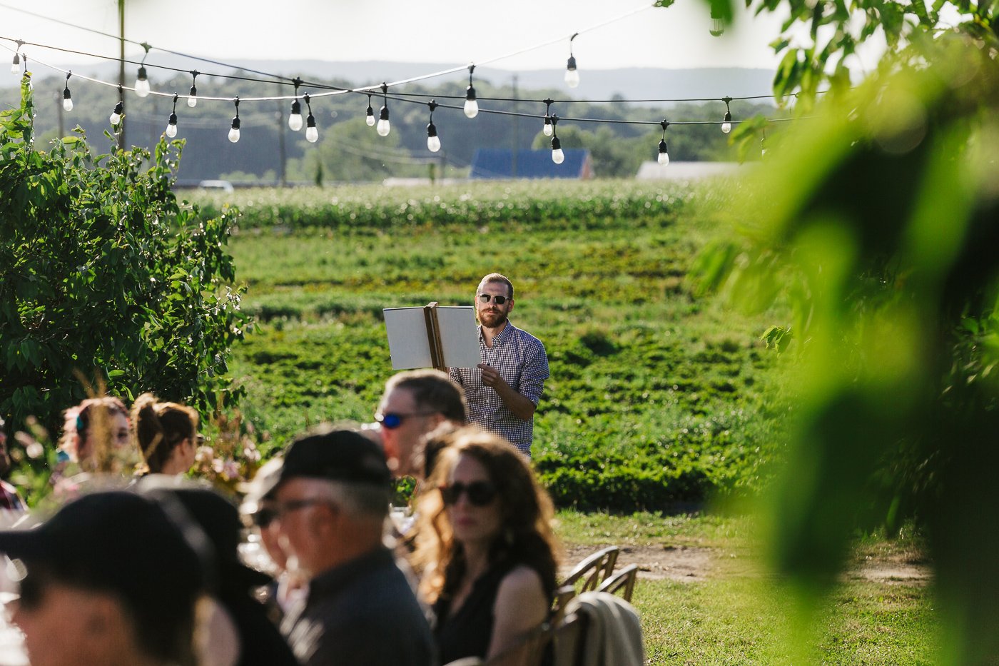 An artist stands outdoors in a scenic field, holding a blank canvas while surrounded by guests at a garden event, with string lights overhead creating a festive atmosphere.