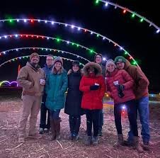 Trail+of+Light+Display+in+PA Group of friends posing together under colorful holiday lights at night, wearing winter coats and hats, enjoying a festive outdoor event.