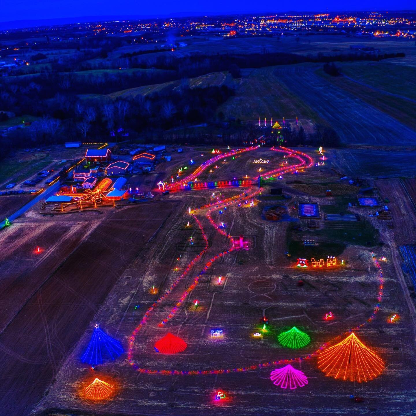 Trail+of+Lights+PA Aerial view of a festive holiday light display featuring colorful illuminated decorations in various shapes, including trees and stars, set against a twilight landscape with a rural backdrop and distant city lights.