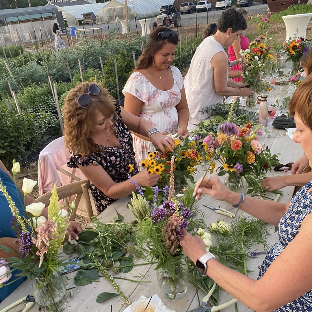 Group of women participating in a flower arranging workshop outdoors, surrounded by vibrant blooms and greenery, with a rustic table set up for crafting floral arrangements.