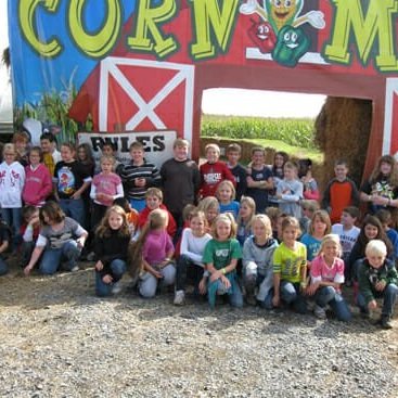 A large group of children gathered in front of a colorful corn maze entrance featuring a barn design, with a sign that reads "Rules." The scene captures a fun outdoor event at a farm, surrounded by cornfields.