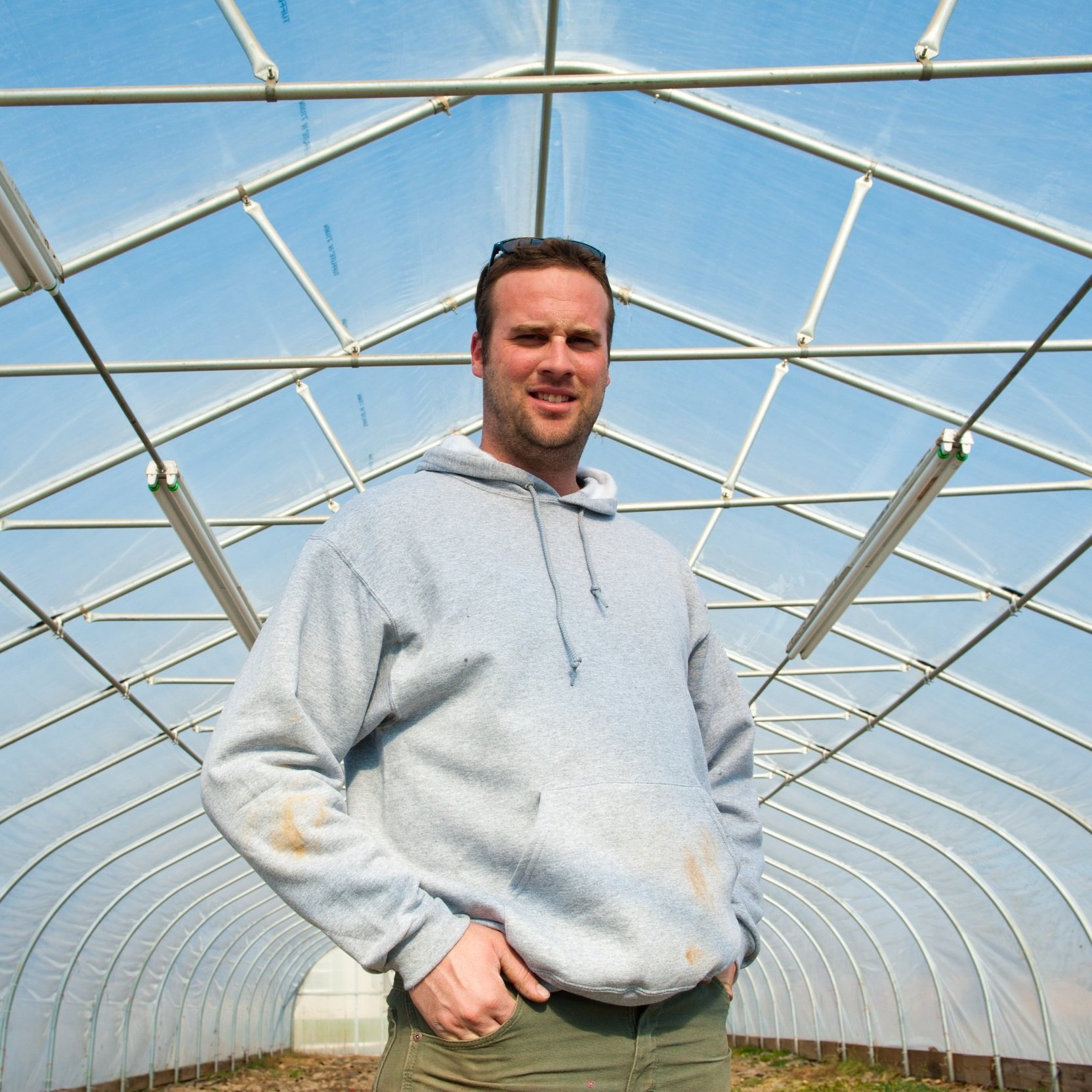 Man standing inside a greenhouse, wearing a gray hoodie and green pants, with a clear blue sky visible through the transparent roof. The structure showcases a modern agricultural environment.