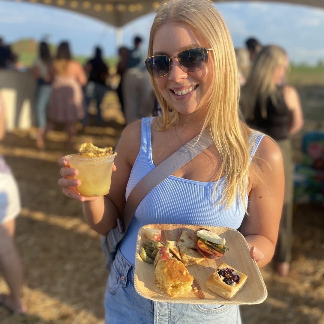 A young woman with long blonde hair and sunglasses smiles while holding a plate of gourmet food and a drink at an outdoor event. The plate features various appetizers, including pastries and vegetables, set against a blurred background of people socializing under a tent.
