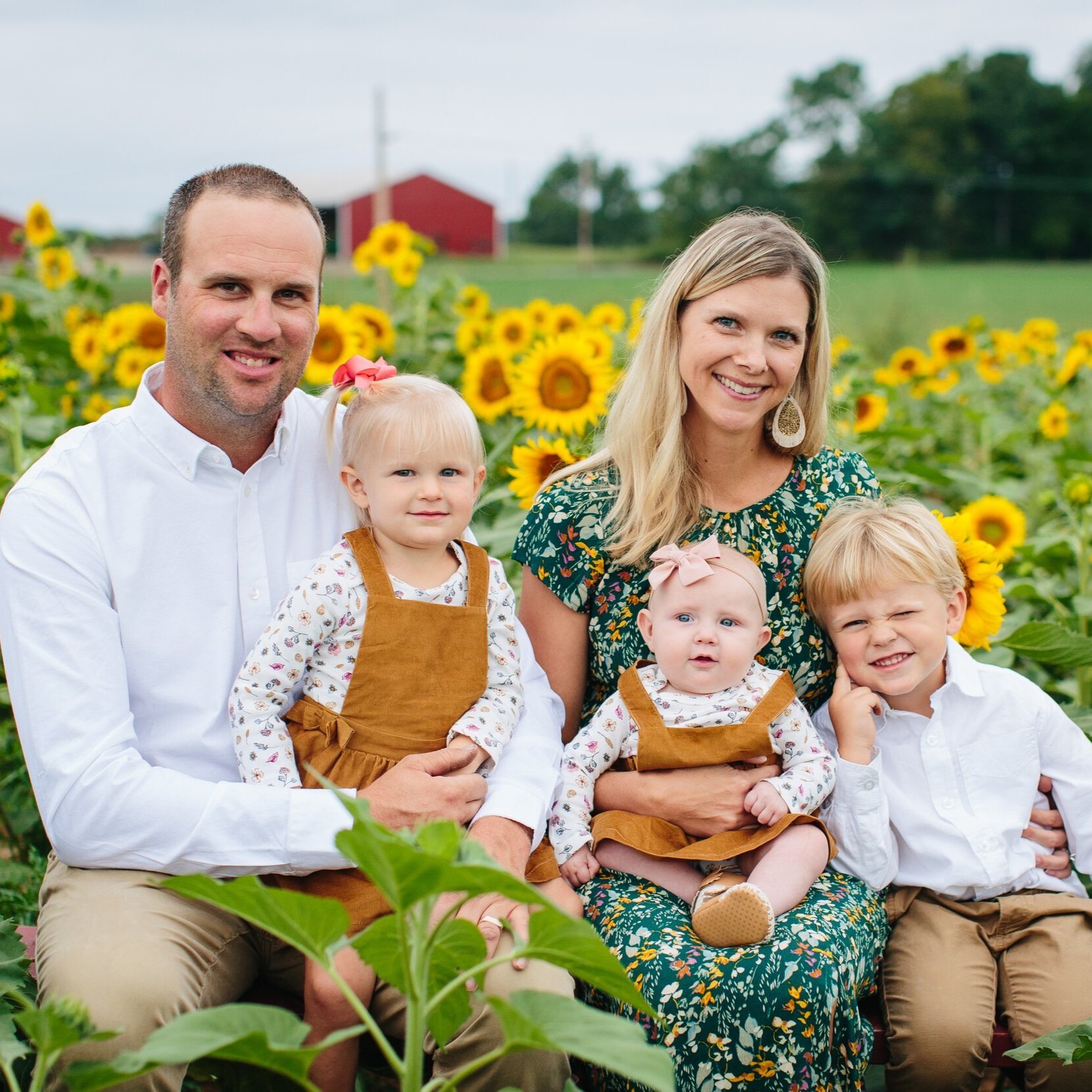untitled-295 (1) Family portrait in a sunflower field, featuring a smiling couple with three young children dressed in coordinating outfits. The background showcases vibrant sunflowers and a rustic barn, capturing a joyful family moment in nature.